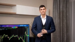 A businessman in a suit stands indoors with a stock market graph on a screen.